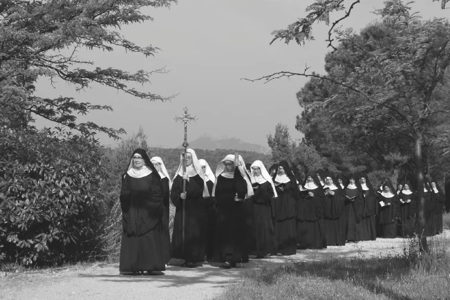 Procession de sœurs religieuses en habit traditionnel noir et blanc, avec une croix processionnelle, marchant sur un chemin de terre extérieur.